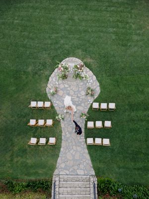 Aerial view of stone pathway leading to wedding ceremony setup with white chairs on green lawn.
