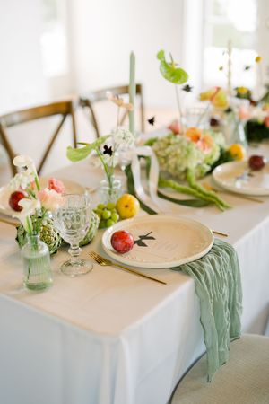 Elegant table setting with soft white linens, mint green accents, fresh flowers in glass vases, and scattered fruits for decoration.