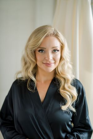 A woman in a black silk robe with long blonde wavy hair poses by a window with natural light.