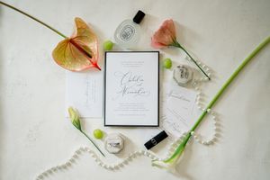 Wedding invitation card styled with anthurium flowers, pearls, and perfume bottles on white surface.