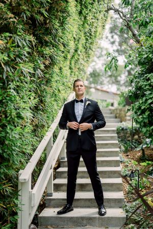 A groom in a classic black tuxedo poses on stone steps surrounded by lush green foliage.