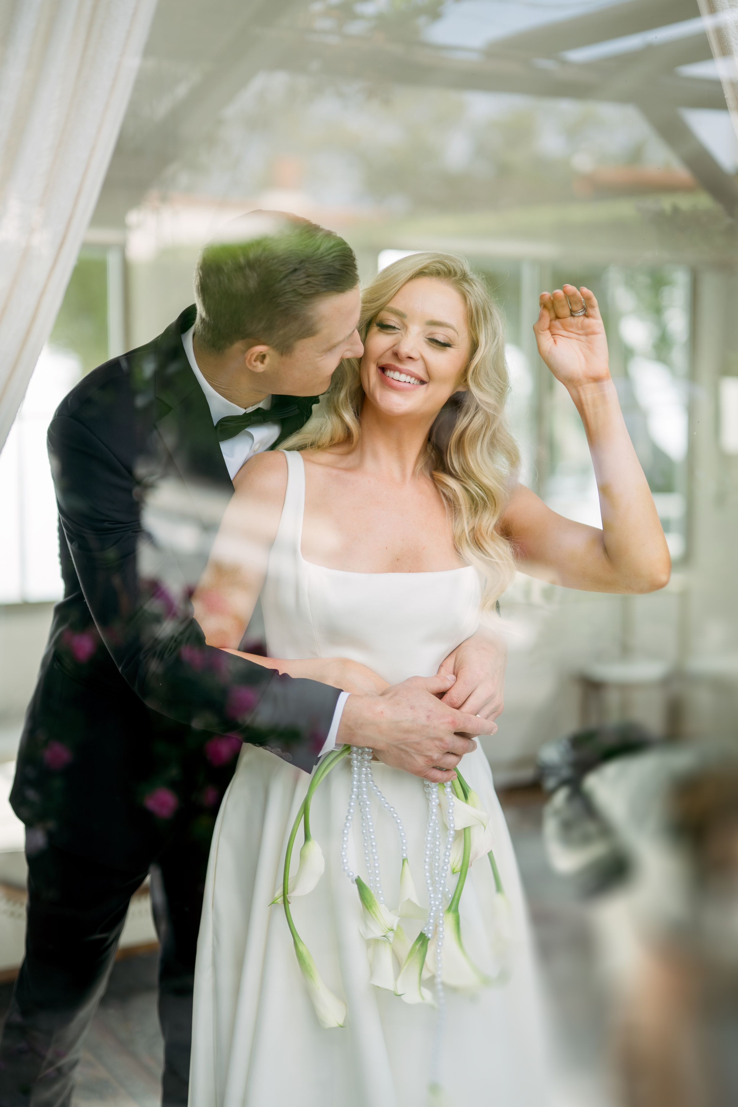 Joyful couple shares an intimate moment during their wedding celebration in a light-filled space with sheer curtains.