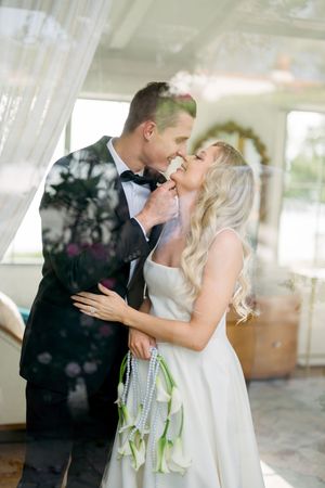 Bride in white dress and groom in tuxedo share an intimate moment under sheer curtains with a wedding bouquet.