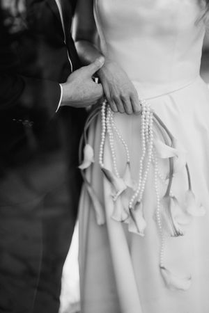 Black and white wedding photo showing hands holding pearl strings and ribbons against a white bridal gown.