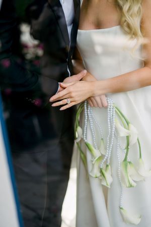 Close-up detail of wedding couple showing white dress, pearl strands and calla lily bouquet with hands clasped together.