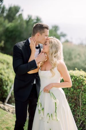 Romantic wedding moment captured outdoors with newlyweds embracing among green foliage and natural scenery.
