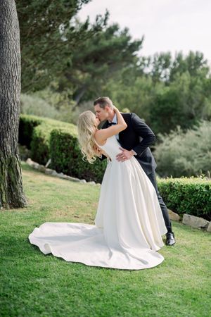 A romantic wedding couple embraces on a lush green lawn with trailing white dress and elegant formal attire.