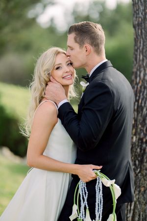 Romantic wedding portrait outdoors with couple sharing tender moment near tree trunk while holding pearl beaded decor.