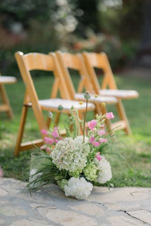 Wooden chairs arranged on a garden lawn with delicate floral arrangements of white hydrangeas and pink flowers lining the stone path.