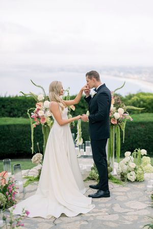 Couple shares intimate moment at outdoor ceremony with floral archway and stone patio surrounded by green landscape and ocean view.