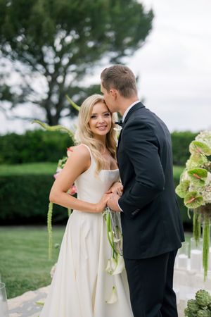 A couple in wedding attire share an intimate moment on a beautiful garden lawn with green trees and floral decor.