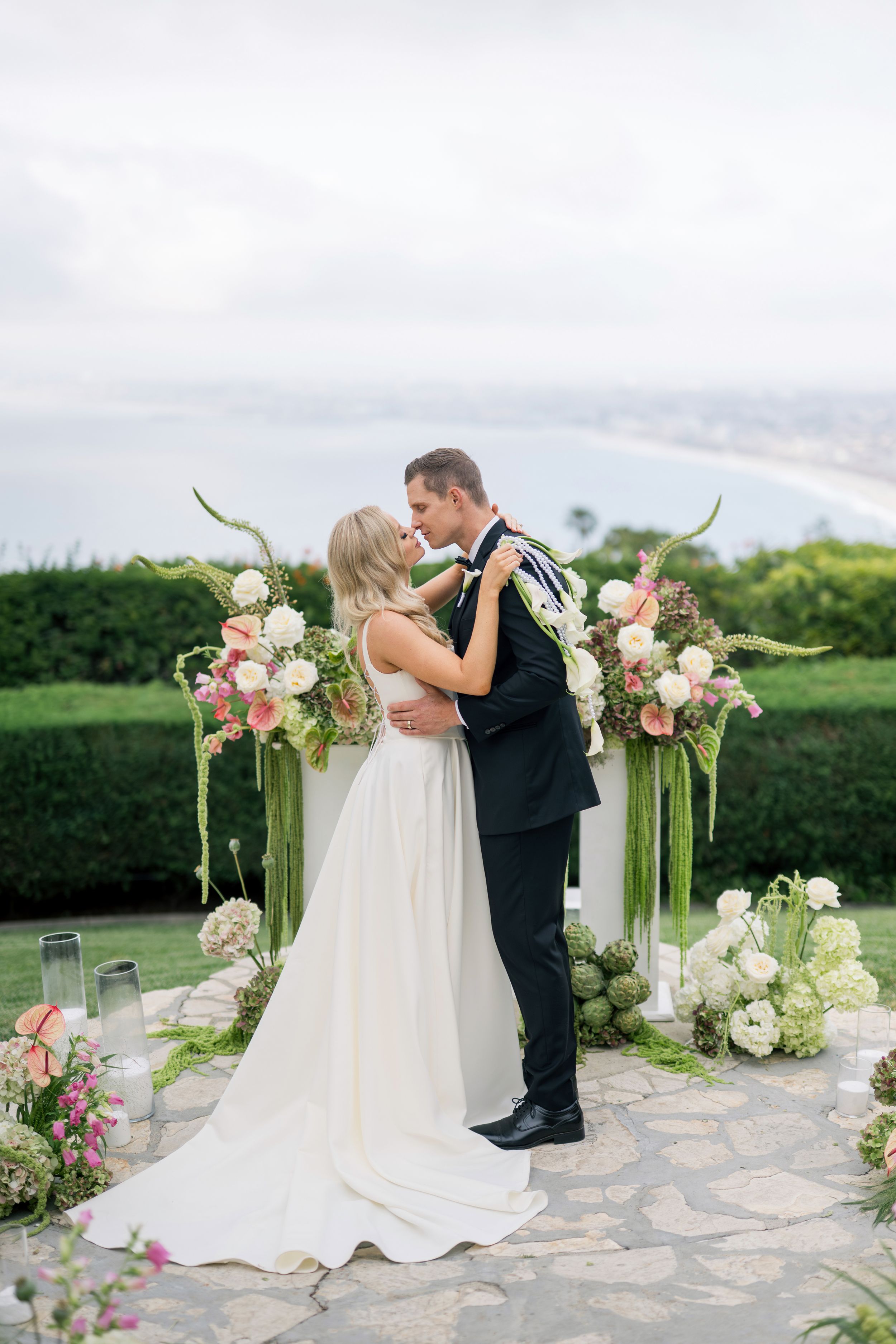 A romantic wedding ceremony arch decorated with pink and white flowers overlooks a scenic coastal view.