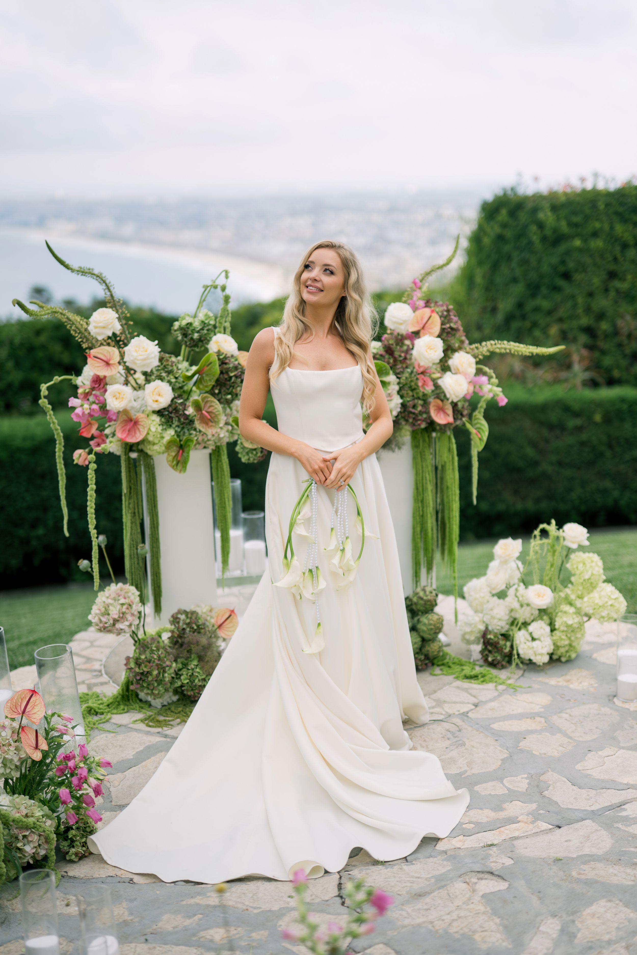 Elegant bride in flowing white gown poses on stone patio with romantic floral arch and seaside backdrop.