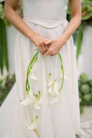 A bride holds an elegant cascading bouquet of white calla lilies with trailing green ribbons against her ivory dress.