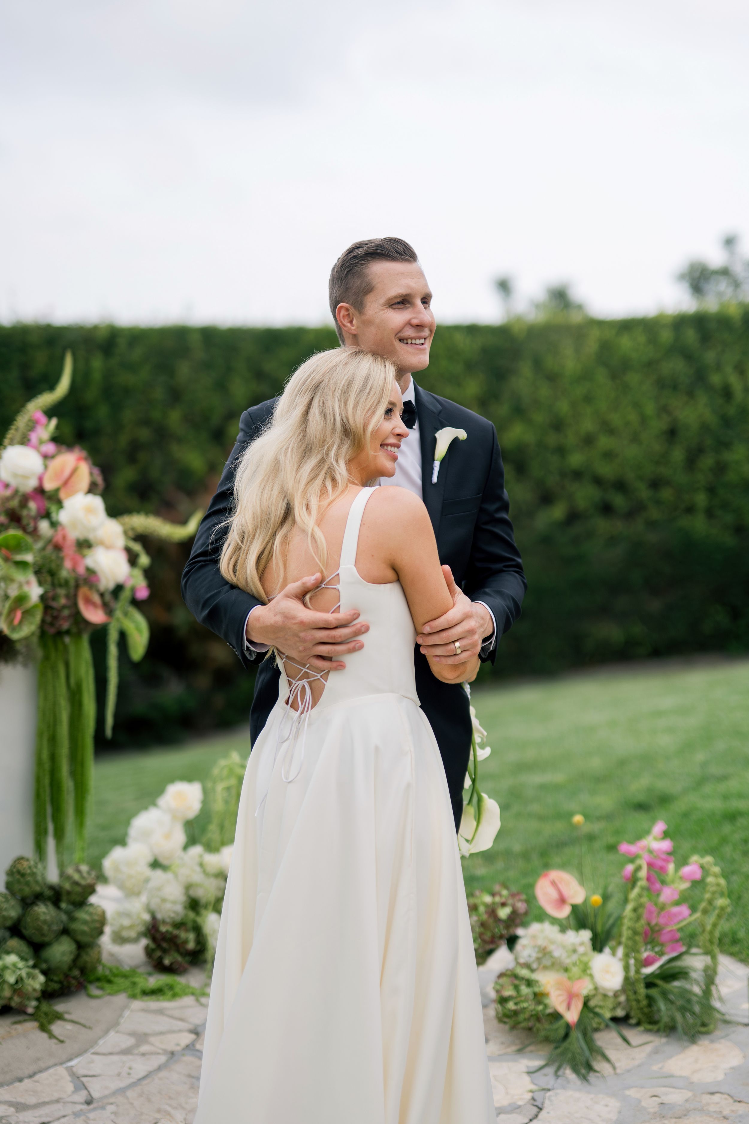 Wedding couple embraces in garden setting with floral arrangements and greenery against stone patio.