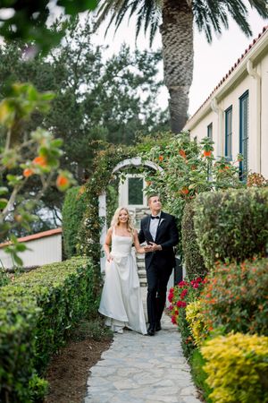 Wedding couple walks along lush garden path with blooming flowers and palm trees at Mediterranean style venue.