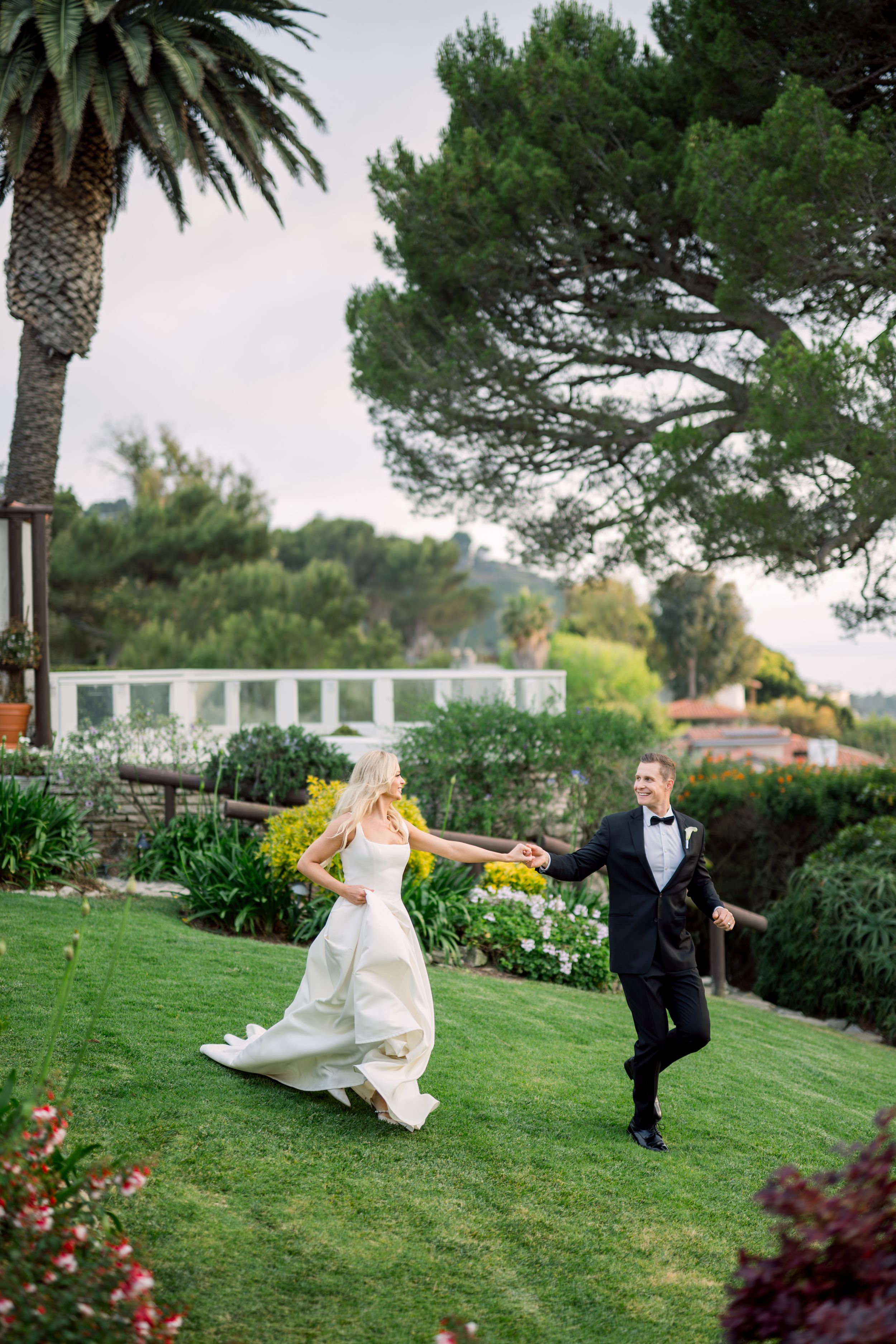 Two people in formal attire run playfully across a lush garden lawn with a palm tree in the background.