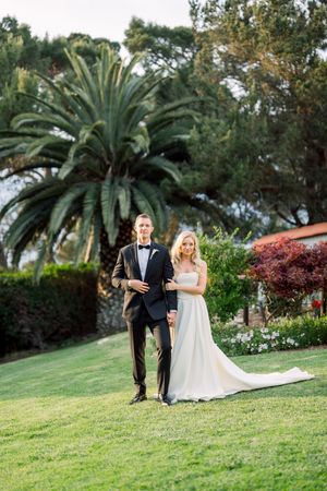 A couple stands together on a manicured lawn with a towering palm tree and colorful landscaping in the background.