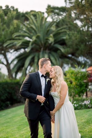 A couple in wedding attire share an intimate moment on a grassy lawn with palm trees and greenery in the background.