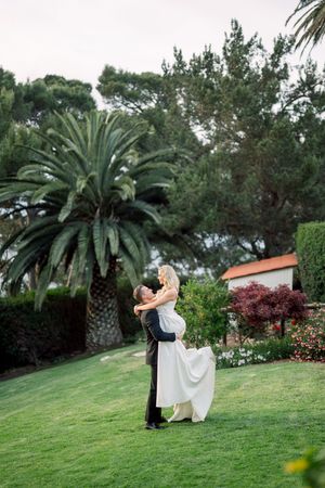 A couple embraces on a lush green lawn with palm trees and garden landscaping in the background.