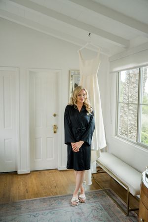 A black silk robe worn before a wedding with a white dress hanging in a bright sunlit room.