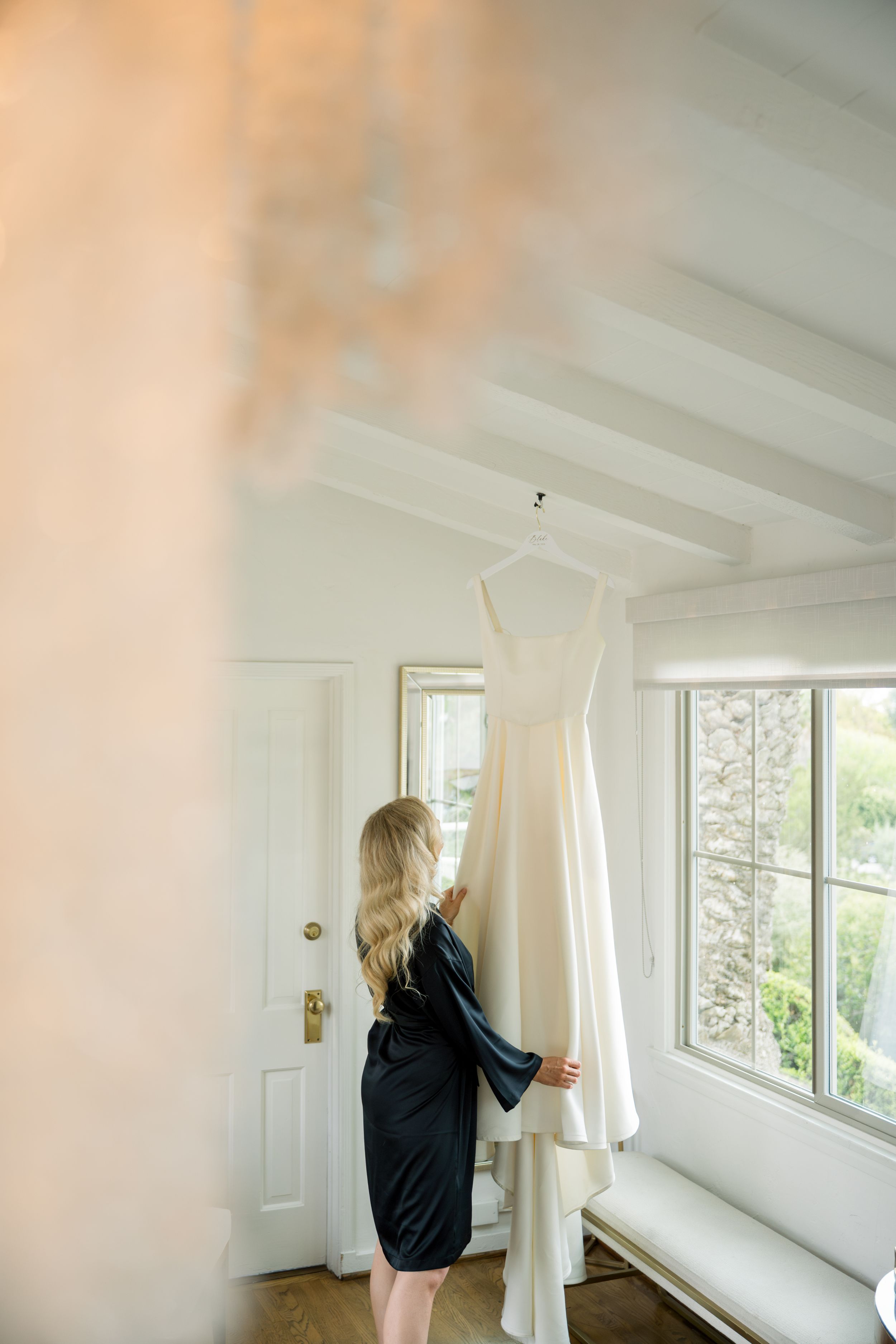 A white wedding dress hangs in a sunlit room while someone in a black robe admires it by a window.