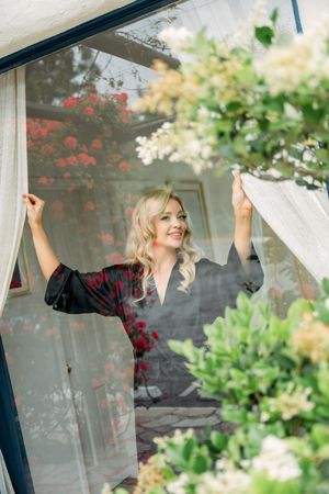 Through a sheer curtain framed by flowering plants, a person in a dark top adjusts window dressings in natural light.