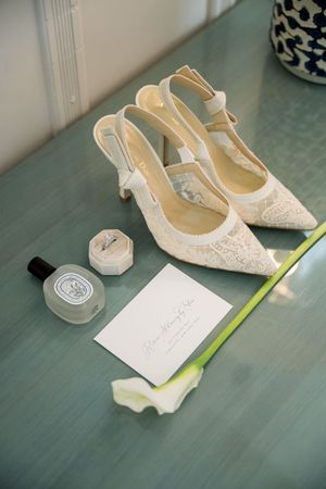 Elegant lace wedding shoes, perfume bottle and note card arranged on turquoise surface.