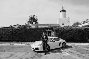 A white Porsche sports car parked in front of a Spanish-style building with a lighthouse tower and palm trees.
