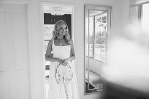 A black and white image shows someone in an elegant white dress standing by a bright window in an interior room.
