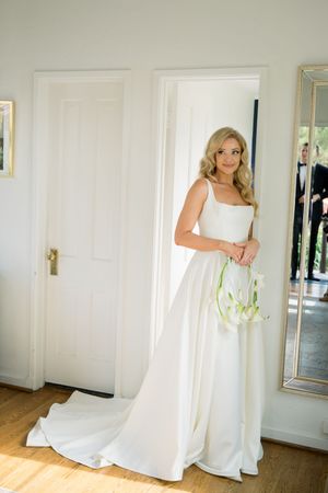 An elegant white wedding gown with a square neckline and flowing train is shown in a sunlit room with white doors.