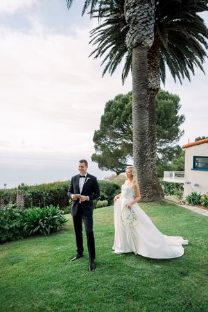 A wedding couple poses outdoors on a lawn with a majestic palm tree and ocean view in the background.