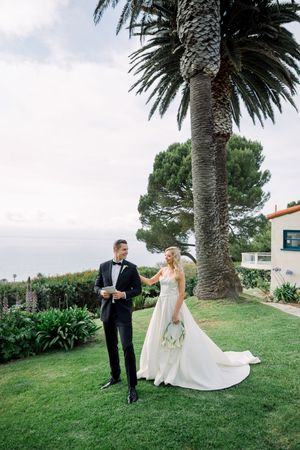 A couple poses by a tall palm tree on a manicured lawn overlooking the ocean on their wedding day.