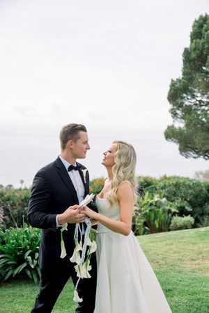 A couple in elegant wedding attire share a joyful moment together on a grassy lawn with greenery in the background.