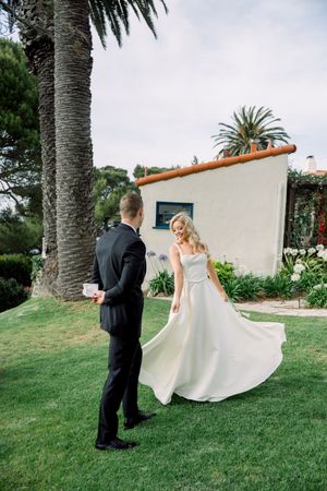 A couple in wedding attire stands on a lawn in front of a Spanish-style building with palm trees in the background.