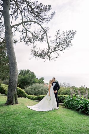 A couple shares an intimate moment on manicured grass under tall pine trees at an elegant outdoor garden wedding.