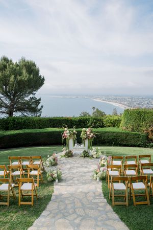 Outdoor wedding ceremony setup with wooden chairs and floral arch overlooking a coastal bay vista on a manicured lawn.