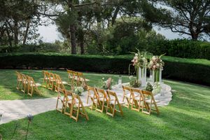 Outdoor wedding ceremony setup with wooden folding chairs and floral arrangements on pedestals in a manicured garden setting.