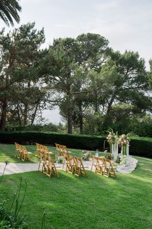 Outdoor wedding ceremony setup with wooden chairs and floral arch against a backdrop of tall pine trees.