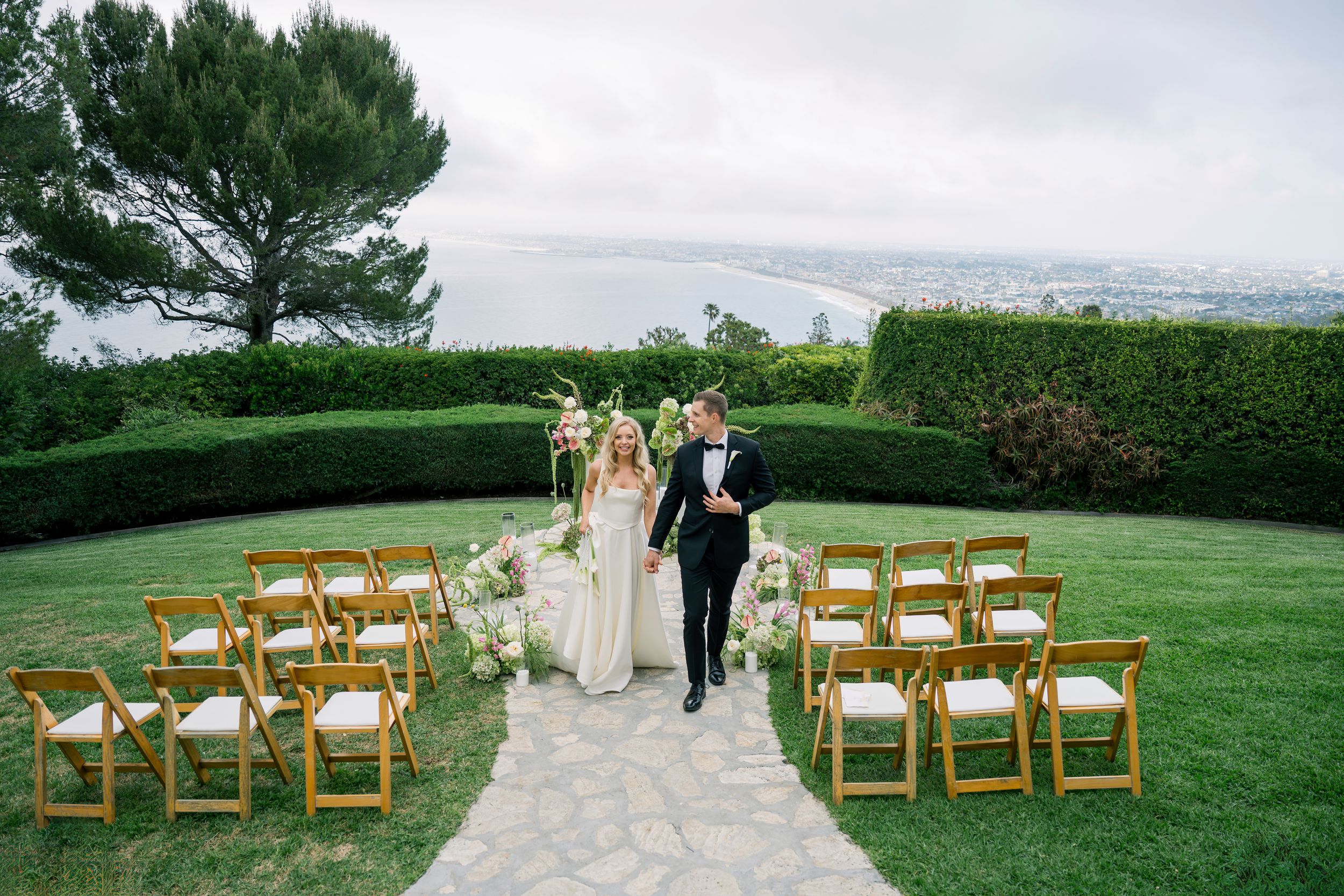 Outdoor wedding ceremony setup on manicured lawn with wooden chairs and stone pathway overlooking coastal view.