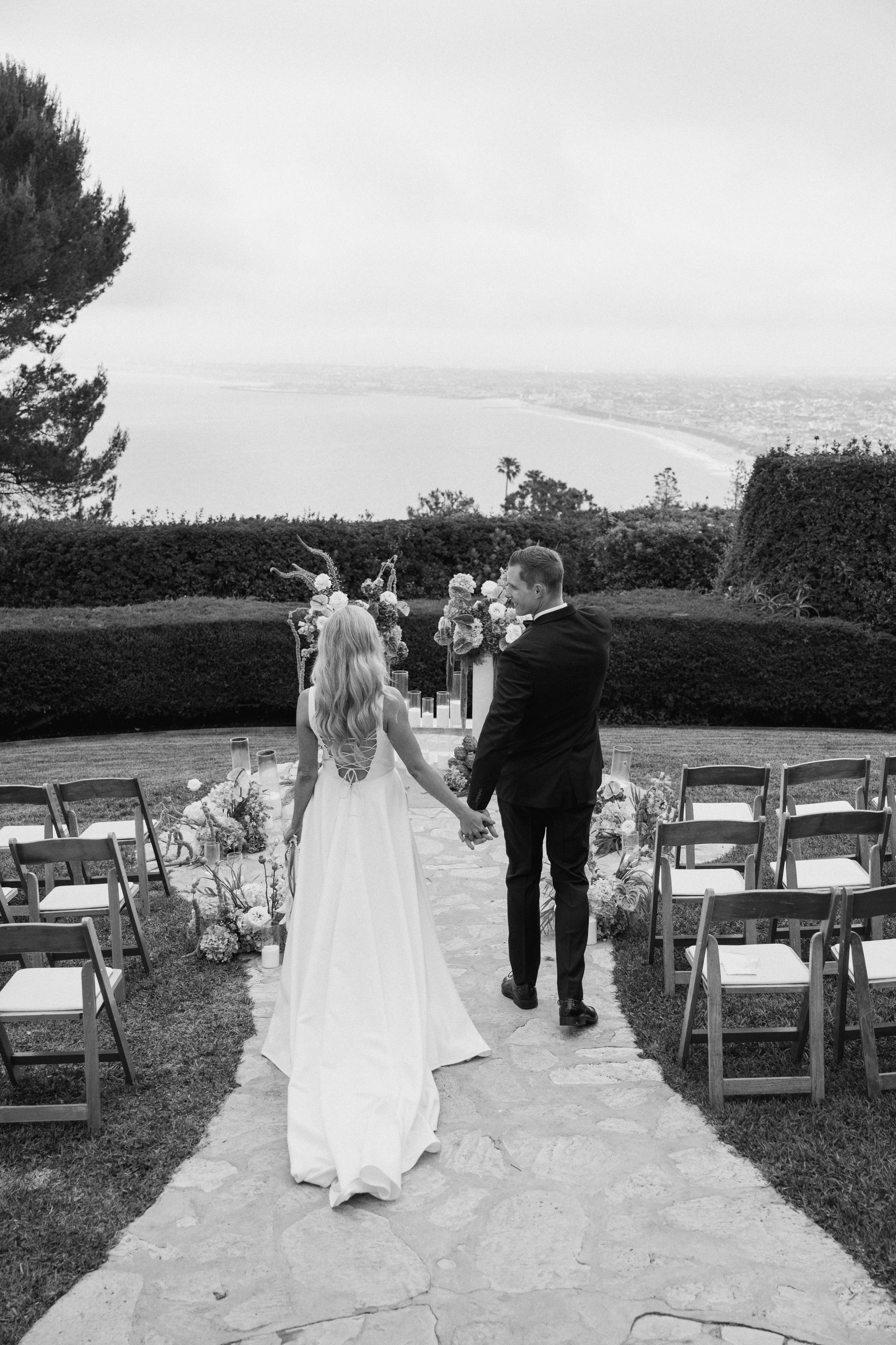 Black and white wedding ceremony photo showing a couple on a stone path with white chairs set up overlooking a coastal view.