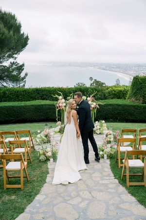 A wedding ceremony setup on a cliffside terrace overlooking a bay, with wooden chairs and floral arch arrangements.