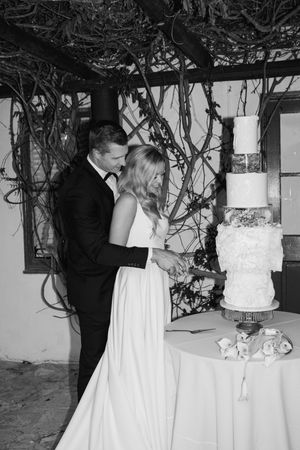 A black and white wedding photo showing a couple cutting their elegant four-tier white cake adorned with vines and greenery.