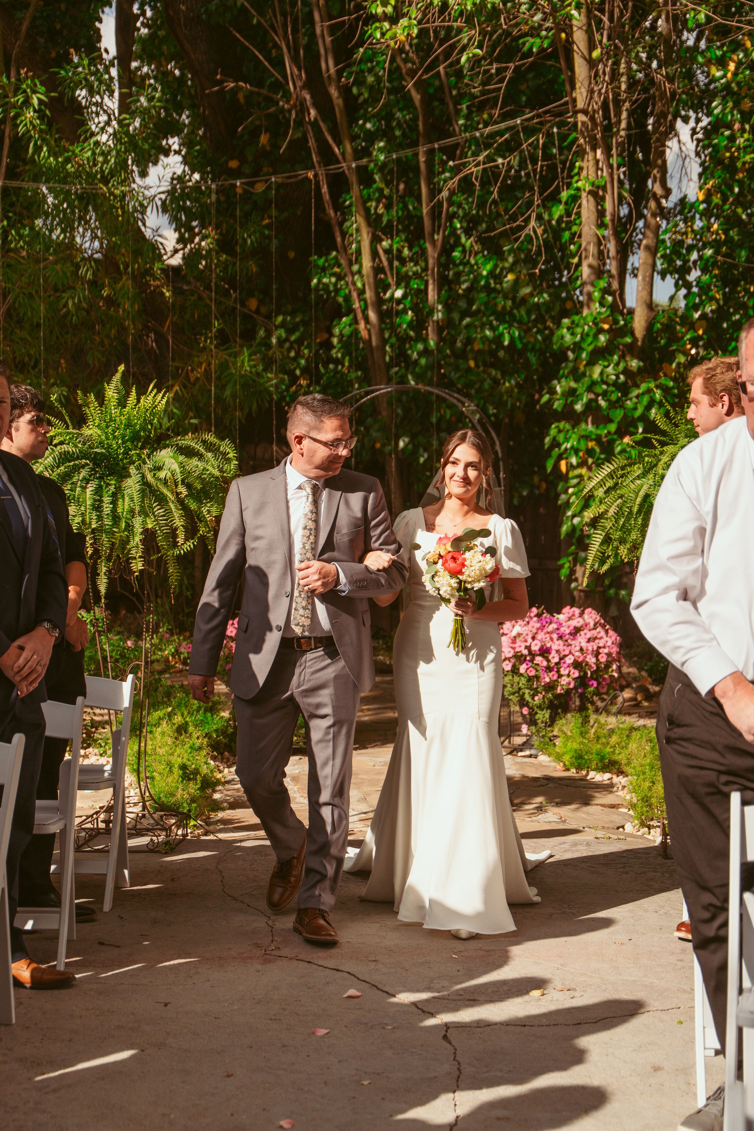 A wedding couple walks down an outdoor garden path lined with lush greenery and ferns.