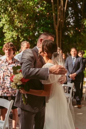 Two people embrace during an outdoor wedding ceremony while guests look on in a sunlit garden setting.