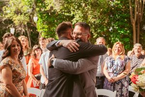 Two grooms embrace during an outdoor wedding ceremony as guests smile and watch from their seats in a garden setting.