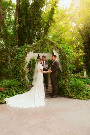 A wedding ceremony takes place under a white arch draped with greenery in a lush garden setting during golden hour.