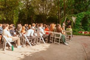 Wedding guests seated in white folding chairs at an outdoor garden ceremony with sunlit natural surroundings.