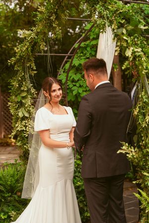 A couple exchanges vows under a beautiful garden arch decorated with lush greenery and white fabric at an outdoor wedding.