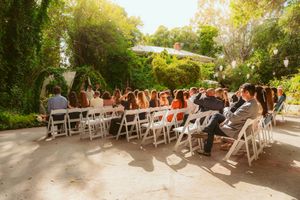 Wedding guests seated in white chairs at an outdoor garden ceremony venue with sunlight filtering through trees.